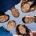 Multi-ethnic group of teenage friends hang out, huddle together with arms around each other outdoors. Blue sky background.