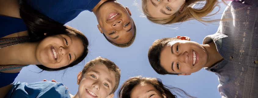 Multi-ethnic group of teenage friends hang out, huddle together with arms around each other outdoors. Blue sky background.